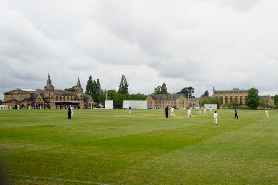 A large, historic cricket ground with well-maintained green outfield and players dressed in white cricket uniforms scattered across the pitch. In the background, there are old stone buildings with poi