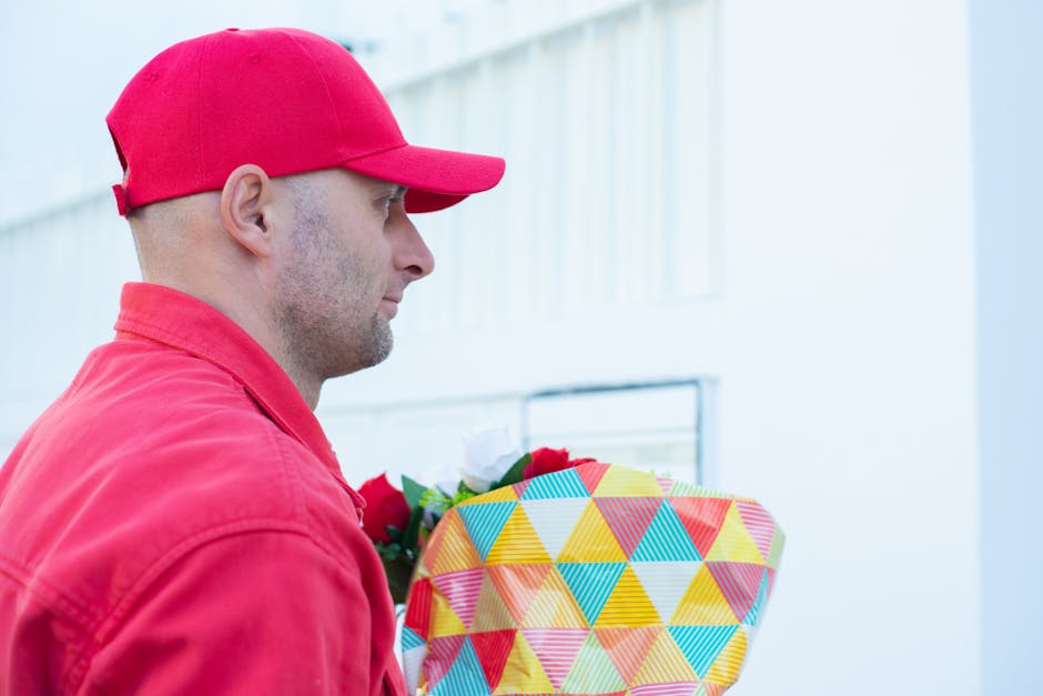 A delivery person wearing a red cap and matching red jacket holding a bouquet of fresh flowers wrapped in colorful geometric-patterned paper with yellow, blue, pink, and green triangles. The bouquet f
