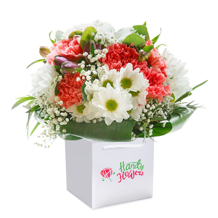 Thoughtfully composed by a skilled florist near St John's Wood High Street, this captivating bouquet sits elegantly in a contemporary white square gift bag, ready to delight. The centrepiece features florid coral and peach carnations, densely layered with signature ruffled petals, radiating both vitality and tenderness. Accentuating them, snow-bright daisies with fresh lime-green centers evoke the joyful charm of local London parks in spring, while sprays of airy white baby's breath weave between the florals, imparting an almost weightless, ethereal touch to the arrangement. On the bouquet's left, glimpses of closed buds, tinged with hints of purple and pink, lend a sense of depth and quiet anticipation-suggestive of new beginnings found walking along Wellington Place. Bold, glossy foliage wraps closely around the base, giving strength and structure, reminiscent of the ancient trees lining St John's Wood's grand roads. The entire floral composition forms a lush, rounded silhouette, its fanned blooms and harmonized greenery highlighted further by the pure white, gently lit background-every detail composed to reflect the elegant sensibilities of the area. White ribbon handles complete the presentation and invite ease of gifting or delivery for birthdays, anniversaries, or welcome gestures throughout St John's Wood. The bouquet stands as a beautiful testament to the freshness, thoughtful selection, and unmistakable quality that local florists bring to every neighbourhood arrangement.