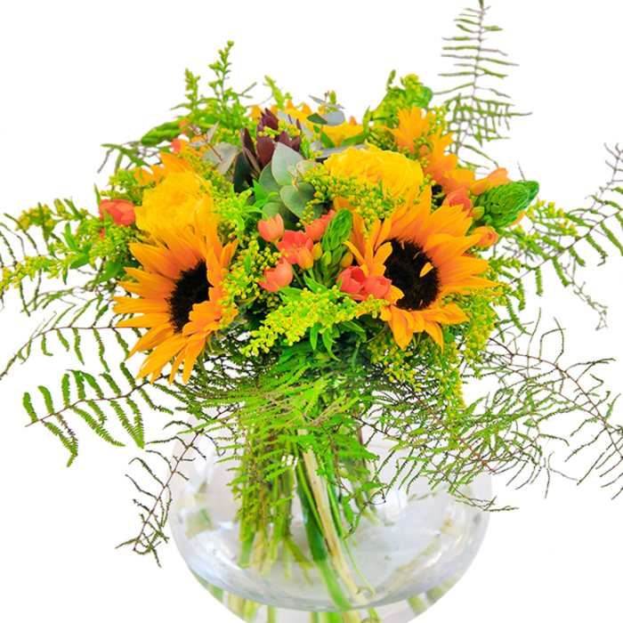 A sun-drenched bouquet brimming with the vivacity of St John's Wood, this artfully composed arrangement captures the very essence of seasonal celebration. Sunflowers with glowing yellow-orange petals and bold, mahogany centers command attention at the core, their shapes reminiscent of sunlight streaming through the canopies of Avenue Road. Alongside, yellow garden roses spiral open with delicate precision, their plush roses exuding a subtle perfume that conjures memories of leisurely strolls in Regent's Park. Ruby-red Hypericum berries are thoughtfully scattered throughout, brightening the composition and offering a joyful contrast to the soft yellows. Cascading layers of feathery ferns, goldenrod sprigs, and slender green foliage envelop the blooms, adding a gently wild silhouette. Hints of cool, bluish eucalyptus complete the air of rustic refinement. The arrangement rests in a perfectly clear, round vase, water refracting the intricate lattice of stems within-an elegant nod to the pure, effortless beauty to be found in St John's Wood's manicured gardens and historic terraces. Every detail, from bold texture to vibrant colour, feels designed to mark life's happy milestones: sending a birthday greeting, welcoming a new arrival, or adorning a summer table for a gathering with friends.